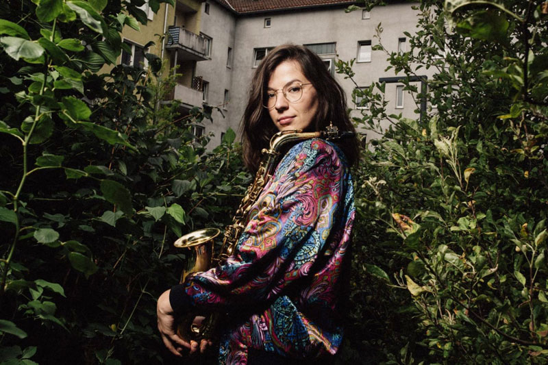 Portraitfoto der Musikerin Theresia Philipp, ein Saxofon haltend. Im Hintergrund ein Gebäude und hohe Sträucher