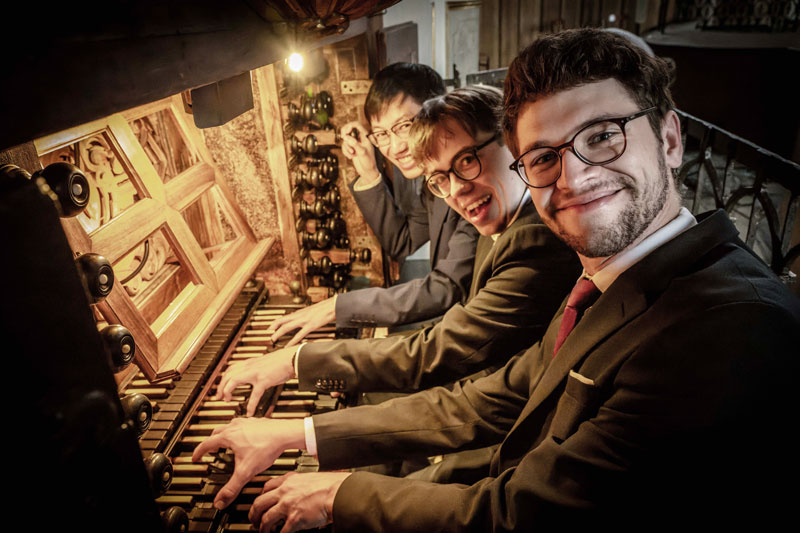 Drei junge Männer sitzen nebeneinander am Spieltisch einer großen Orgel und schauen mit glücklichem Gesichtsausdruck in Richtung Kamera