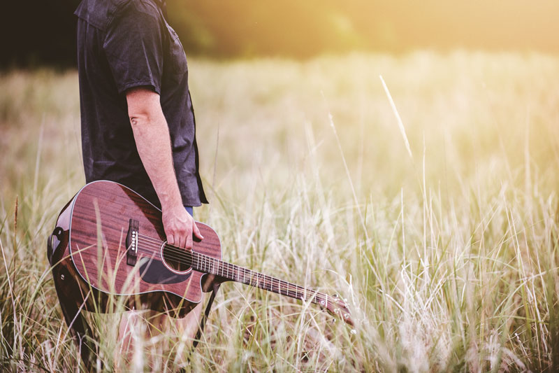 Mann, dessen Kopf man nicht sieht, mit Gitarre in der rechten Hand in einem sommerlichen Kornfeld bei romantischem Gegenlicht