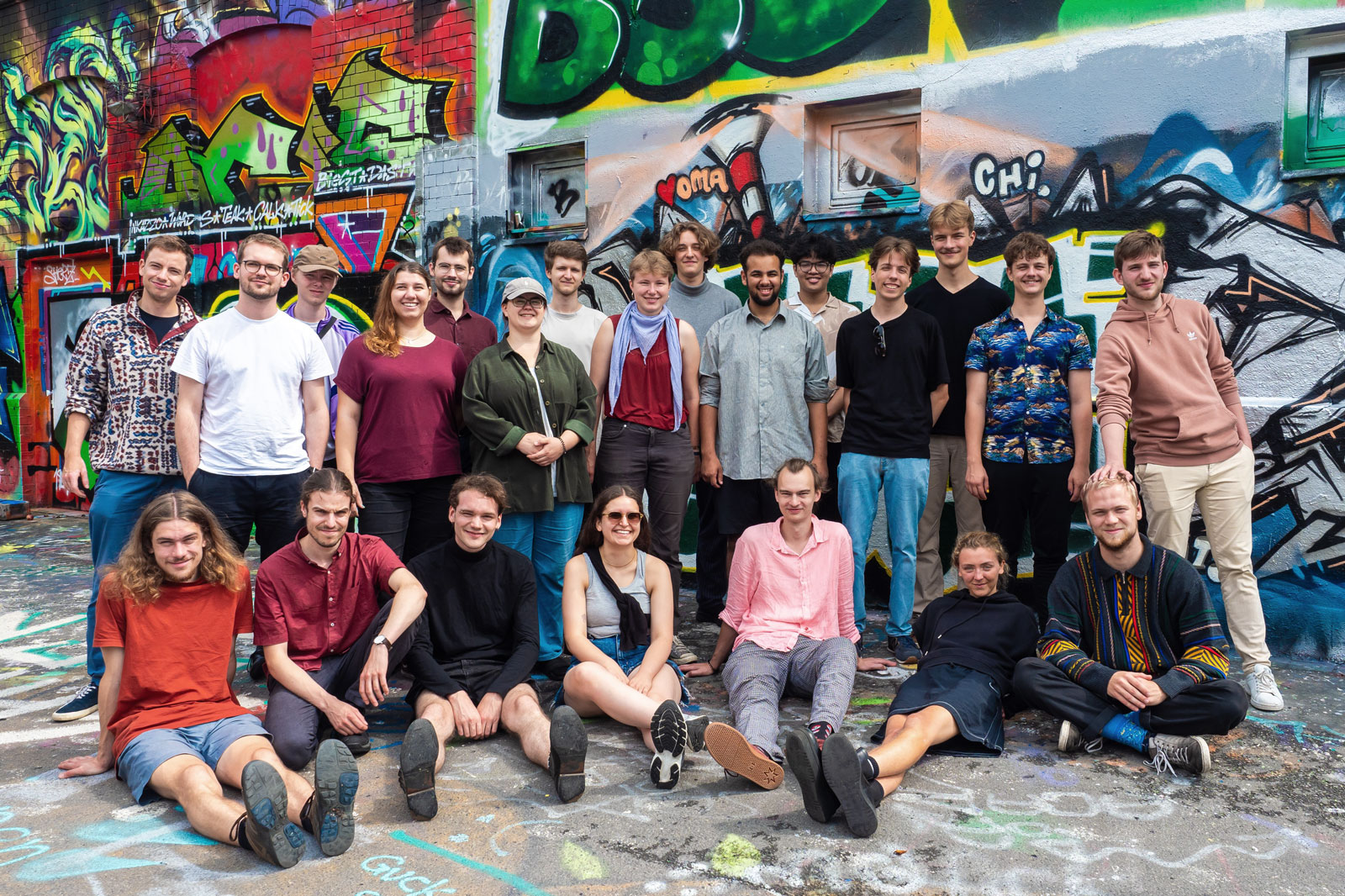 Gruppenfoto mit jungen Menschen, einige im Vordergrund auf derm Boden sitzend, die anderen dahinter stehend. Im Hintergrund eine mit Graffiti besprühte Wand