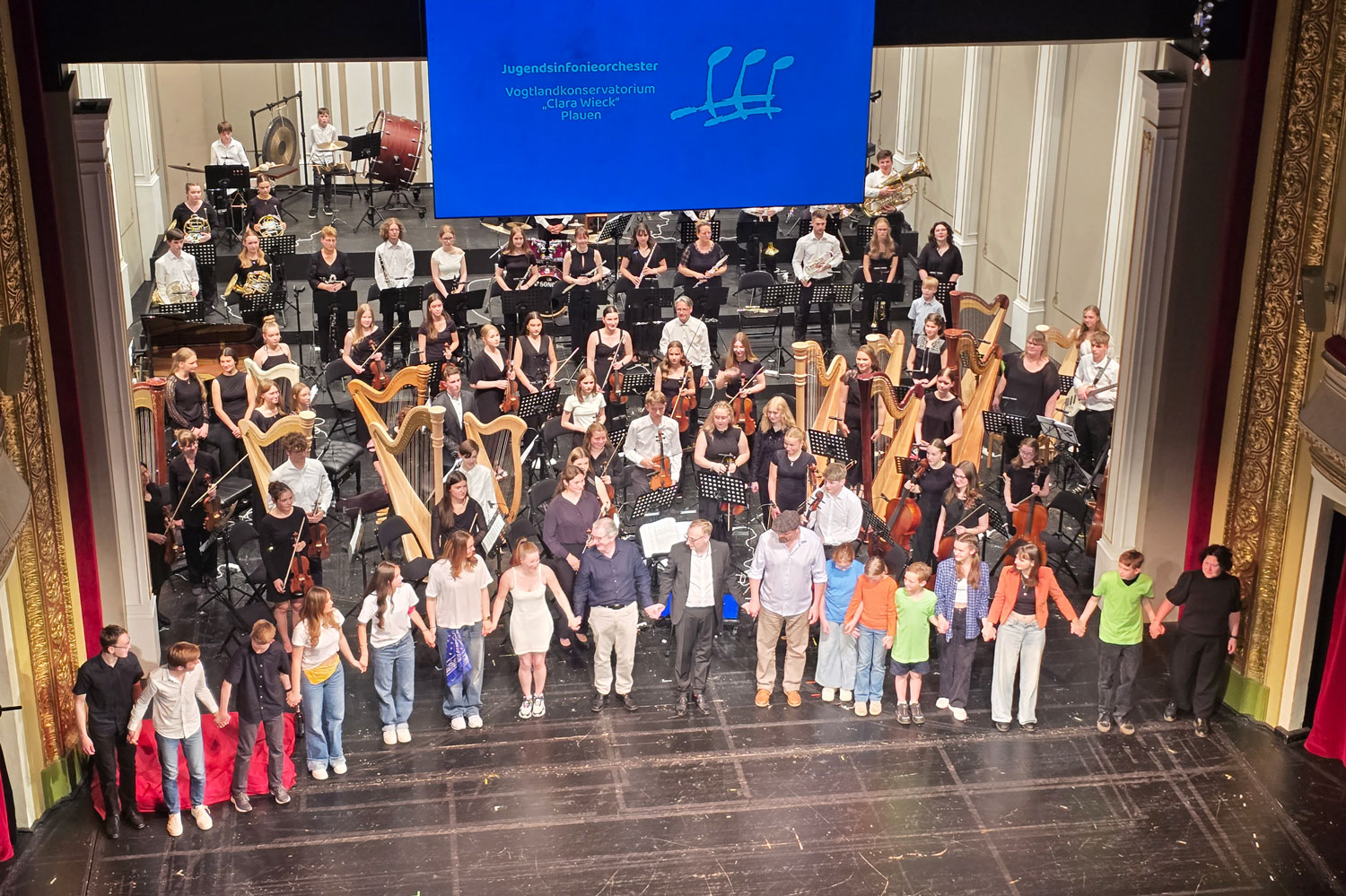 Viele Menschen mit und ohne Instrumente stehen auf der Bühne des Theaters Plauen. Das Foto wurde vom Rang aus aufgenommen.