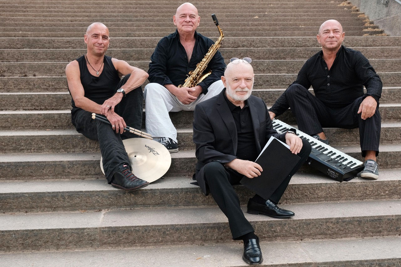 Küf Kaufmann und das Jerusalem Trio: Vier kahlpöpfige Herren, überwiegend schwarz gekleidet sitzen auf eine Steintreppe. Sie halten Schlagzeugsticks, Sxophon, Notenmappe und Keyboard jeweils in ihren Händen