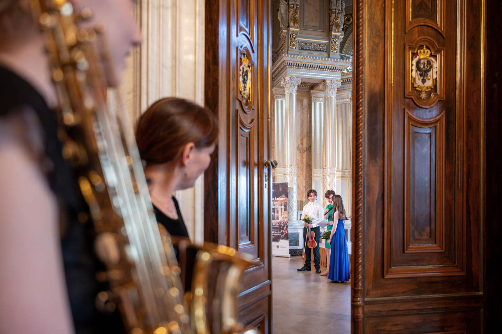 Durch eine geöffnete Tür blickt man in den Kronensaal des Schlosses Albrechtsberg. Dort stehen drei Jugendliche mit Instrumenten und Blumen in der Hand nach ihrem Auftritt. Im Vordergrund sind, noch vor der Tür stehend, zwei Jugendliche zu erkennen, die gleich durch diese Tür zu ihrem Auftritt gehen werden.