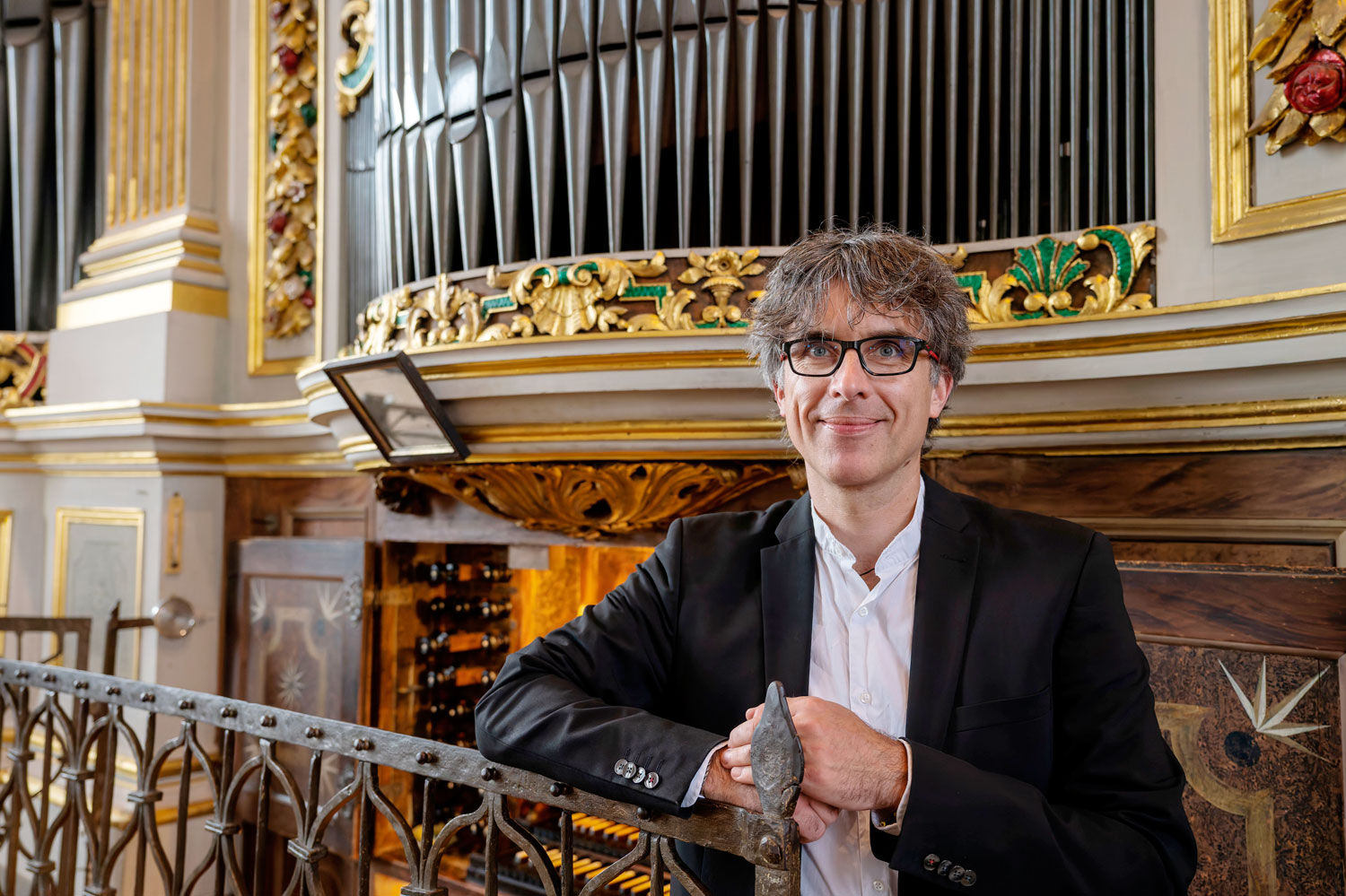Ein freundlich in die Kamera blickender Mann mit Brille. Im Hintergrund eine Orgel